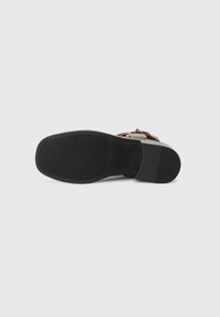 Sandale marron avec une semelle en caoutchouc noir, présentant un design plat, une surface texturée et un accent de boucle en ton bronze.