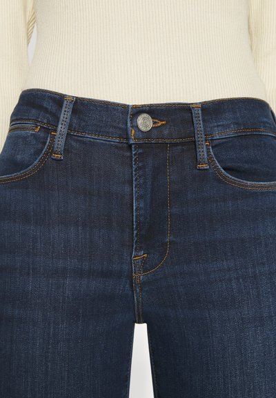 Jean en denim bleu foncé au design ajusté. Comprend une fermeture à bouton à l'avant, des coutures jaunes et des poches latérales.