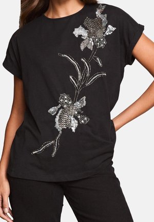 Haut noir à manches courtes avec un motif floral en sequins argentés sur le devant, porté avec un pantalon noir assorti.