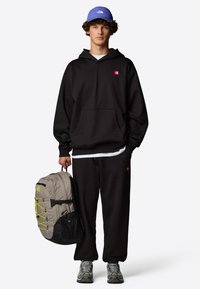 Czarna bluza z kapturem z czerwonym logo, czarne dresy, szary plecak z neonowymi akcentami i szare buty. Czapka jest niebieska z logo.