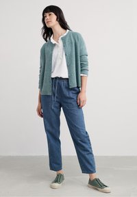 Kvinne iført teal cardigan over hvit bluse, blå denimbukser med frontlommer, og grønne sneakers, stående mot en enkel lys bakgrunn.