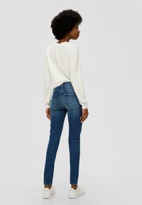 Bijela teksturirana majica dugih rukava u kombinaciji s plavim skinny trapericama. Bijele tenisice upotpunjuju outfita. Pogledano odostraga.