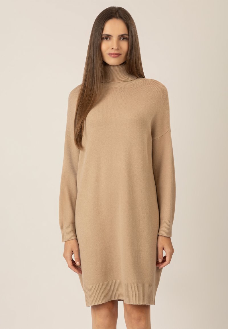 Beiges Strickkleid mit hohem Kragen, tief sitzenden Schultern und langen Ärmeln. Mit gerippten Akzenten an den Bündchen und dem Saum. Weicher Stoff.