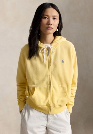 Jeune femme aux longs cheveux foncés portant un sweat à capuche zippé jaune clair avec cordons et un pantalon blanc, debout les mains dans les poches.