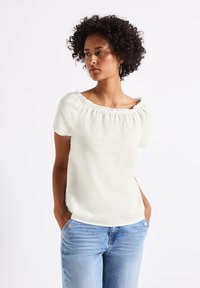 Top corto a maniche corte di colore crema chiaro con scollatura arricciata off-the-shoulder, realizzato in tessuto testurizzato, abbinato a jeans blu chiaro.