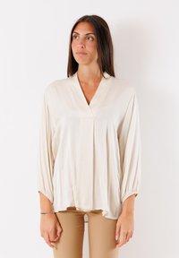 Blusa beige avvolgente con maniche a sbuffo lunghe, texture morbida e un leggero lucido; presenta un collo a V e polsini arricciati. Indossata con pantaloni chiari.