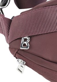 Fialová látková taška s texturovaným popruhem, stříbrnou kováním a táhlem na zip s detailem loga 'B'. Kompaktní a funkční design.