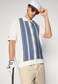 Giovane con berretto bianco, occhiali da sole e maglione a righe con bottoni, che tiene una mazza da golf con guanto bianco su uno sfondo semplice.