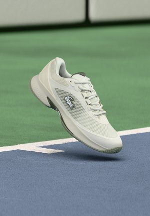 Valkoinen urheilukenkä, jossa krokotiililogo, leijuu tenniskentän viivan yllä sinivihreällä pinnalla.