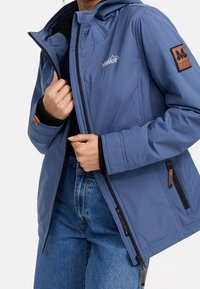 Osoba ve modré bundě s logem "Mountain", černé vnitřní vrstvě a modrých džínách, upravuje zip na přední části bundy.