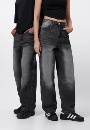 Deux personnes portant des jeans noirs délavés à jambes larges et des hauts noirs, debout côte à côte, les mains dans les poches, devant un fond uni.
