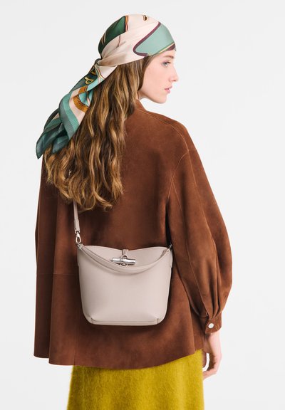 Beige leather crossbody bag with a structured shape, featuring a silver clasp and a short strap, worn over a brown suede jacket and green knit skirt.