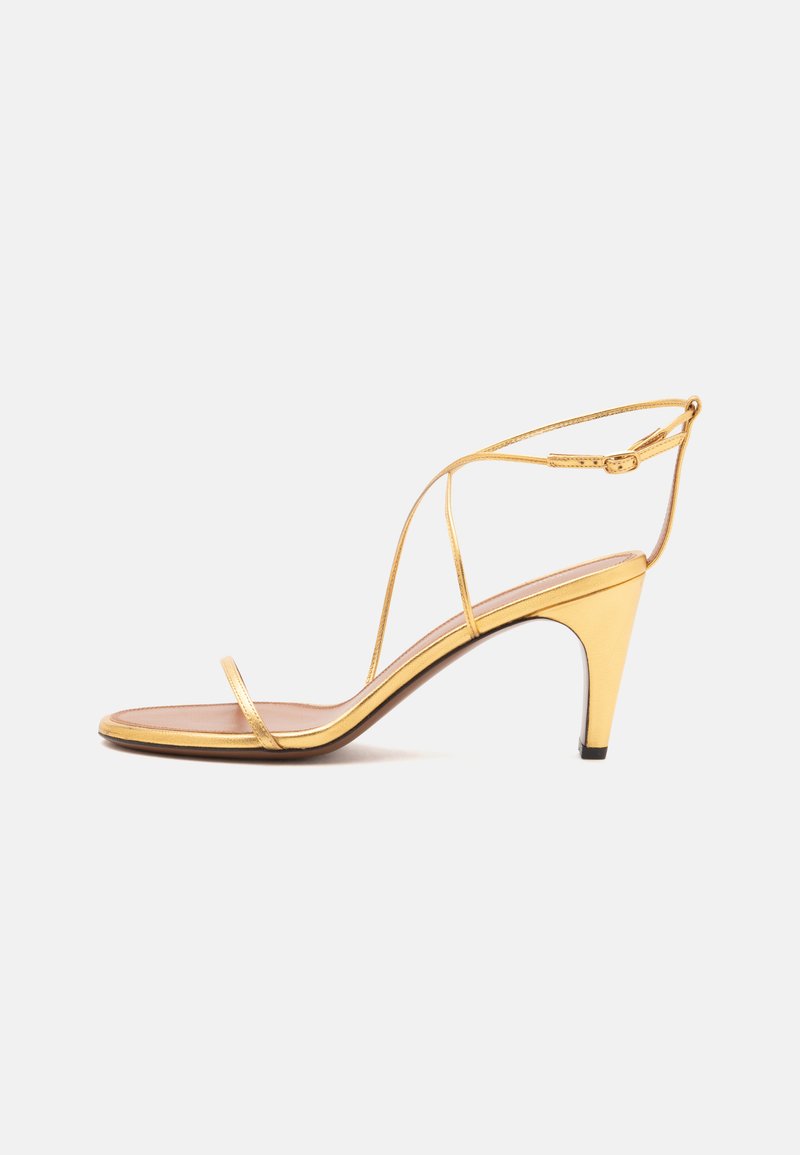 Sandale à talons hauts dorée avec fines lanières et une fine boucle à la cheville sur un fond gris clair.