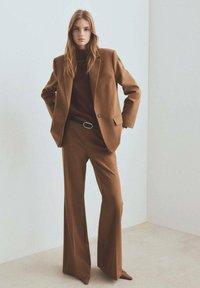 Chaqueta marrón a medida y pantalones de pierna ancha a juego, con una blusa marrón ajustada debajo, acentuada con un cinturón azul y zapatos marrones de punta.