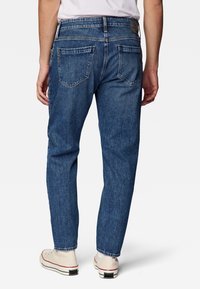 Mies joka käyttää sinisiä farkkuja ja valkoisia tennarikenkiä, kuvattu takaa yksinkertaista valkoista taustaa vasten.