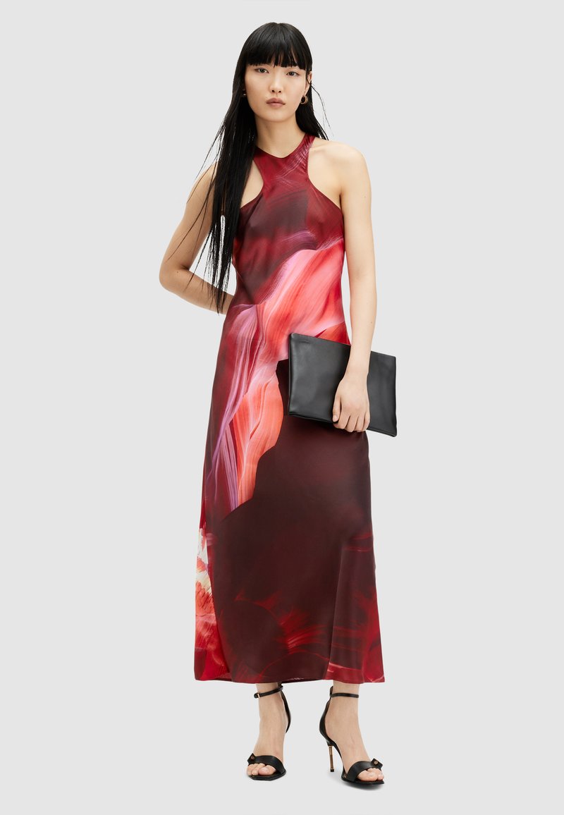 Maxi dress in red and pink abstract print, halter neckline, fitted bodice, flowing fabric. Model holds a black clutch and wears black heels.