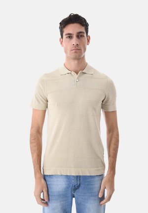 Uomo con polo beige a maniche corte e jeans azzurri, in piedi fronte alla fotocamera su uno sfondo bianco semplice.