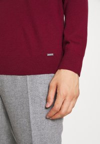 Pull bordeaux en tissu doux, avec un ourlet côtelé et une petite étiquette logo en argent. Un pantalon gris texturé est partiellement visible.