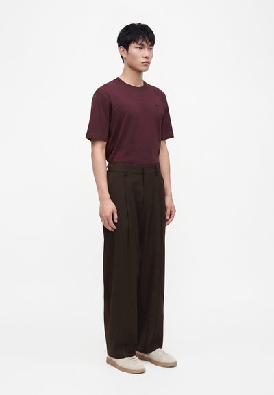 Jeune homme portant un t-shirt bordeaux, un pantalon large foncé et des baskets beiges, debout devant un fond clair uni.