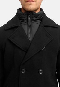 Schwarzer doppelt knopfbarer Wollmantel mit großen Revers und zwei schwarzen Knöpfen, über einer hochkragen schwarz gefütterten Jacke geschichtet.