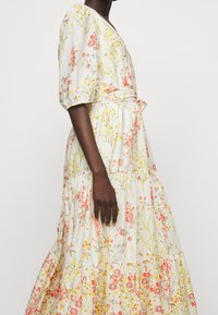 Blommig maxiklänning i cream med rosa och gula mönster, korta puffärmar, v-ringning och ett knytband vid midjan. Tillverkad av lättviktsmaterial.