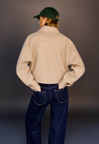 Beige woolen cropped jacket with a collared neckline and wide sleeves, paired with dark blue denim jeans featuring contrast stitching. Green cap accent.