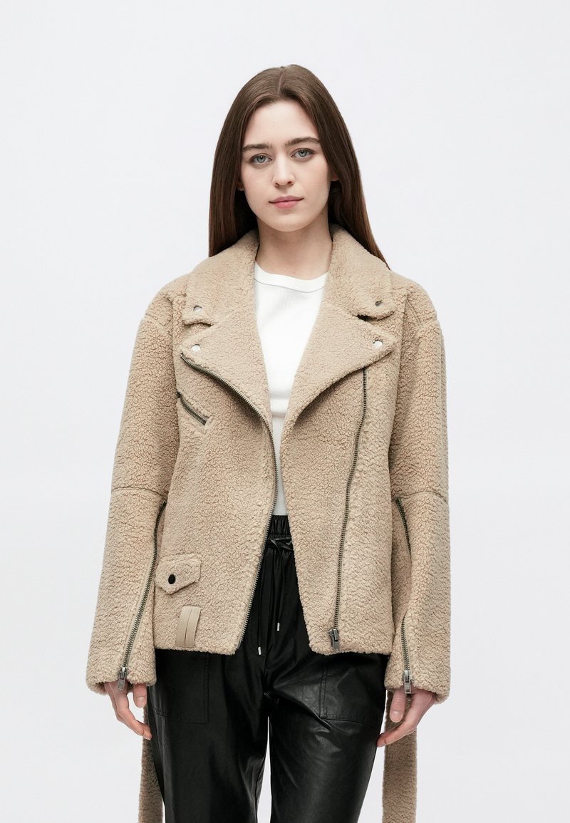 Jonge vrouw die een beige sherpa-jas met ritsen en zwarte leren broek draagt, staand tegen een effen witte achtergrond.