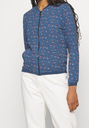 Femme portant une veste bleue zippée avec de petits motifs floraux roses et blancs et un pantalon blanc, debout devant un fond uni.