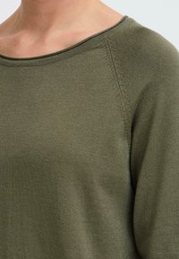 Olíva zöld pulóver kerek nyakkivágással és strukturált varrással a vállaknál. Puha anyag sima felülettel. Közeli nézet az anyagról.