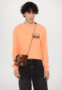 Neon orange langærmet skjorte med "Aries" grafik; sorte højtaljede jeans; brun læder crossbody taske med pletmønster og kædestrop.