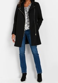 Černý kabát se strukturovaným designem, obsahující zapínání na zip, boční kapsy a hladkou texturu. Nositelný přes vzorované tričko a modré džíny.
