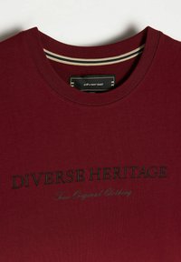 Bordowa koszulka z okrągłym dekoltem, z haftem "Różnorodna Dziedzictwo" w ciemnozielonym kolorze oraz metką z napisem "różnorodne" przy kołnierzyku.