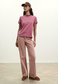 Young woman wearing a mauve t-shirt, light pink wide-leg jeans, brown sneakers, purple necklace, and a dark brown cap standing on a gray floor.