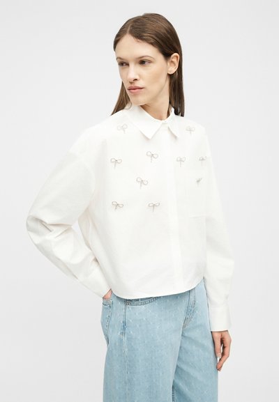 Γυναίκα με ίσια καστανά μαλλιά που φοράει λευκό πουκάμισο με κουμπιά και ασημένιες διακοσμήσεις σε σχήμα φιόγκου, καθώς και ανοιχτόχρωμα τζιν, κοιτάζει αριστερά με το χέρι στην τσέπη.