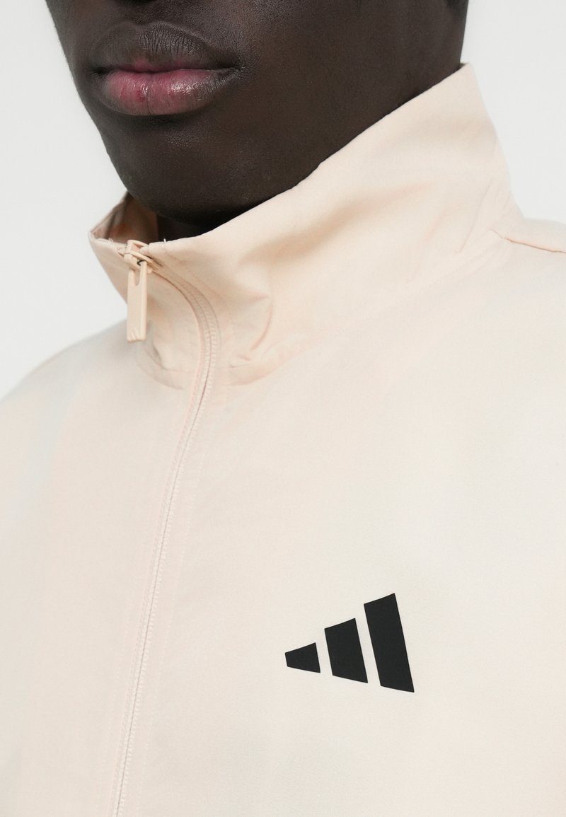 Veste à fermeture éclair de couleur pêche avec un col montant et un logo géométrique noir sur la poitrine. Confectionnée en tissu lisse avec une légère brillance.