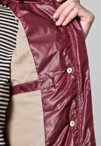 Fodera imbottita color bordeaux con texture liscia, con uno strato interno beige, bottoni a pressione e tessuto a righe a contrasto.