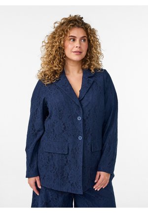 Frau mit lockigem Haar, die ein marineblaues Spitzenblazer mit Knöpfen und passende Hose trägt und leicht zur Seite blickt vor weißem Hintergrund.