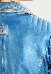 Nahaufnahme einer Person, die eine blaue Lederjacke mit perforiertem Kragen und einem metallenen Markenlogo-Patch auf dem Rücken trägt.