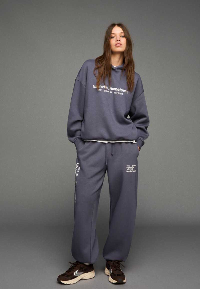 Donna che indossa un ampio hoodie grigio e pantaloni della tuta abbinati con testo bianco, in piedi con le mani nelle tasche e scarpe da ginnastica marroni su uno sfondo grigio.