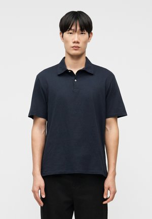 Mies seisomassa kasvot eteenpäin, yllään tummansininen lyhythihainen poolopaita ja mustat housut, yksinkertaista vaaleaa taustaa vasten.