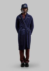 Marineblauwe badjas van badstof met witte piping, voorzien van een voorzak. Gepaard met burgundy geprinte broek en zwarte schoenen.