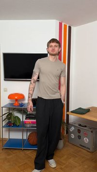 Joven con tatuajes que lleva una camisa beige y pantalones negros, de pie en una habitación con un balón de baloncesto, una lámpara naranja y una maleta con pegatinas.