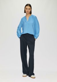 Lichtblauw, langek mouw shirt met een V-hals en geplooide schouders, gecombineerd met donkere, rechte broek en lichtgekleurde schoenen.
