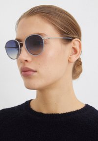 Lunettes de soleil rondes avec des verres dégradés bleu foncé, monture en métal argenté et détails subtils sur les branches, portées par un mannequin avec un pull sombre.