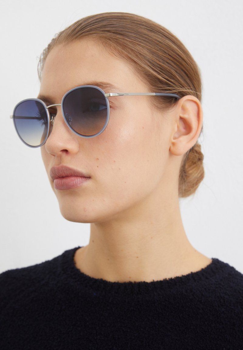 Lunettes de soleil rondes avec des verres dégradés bleu foncé, monture en métal argenté et détails subtils sur les branches, portées par un mannequin avec un pull sombre.