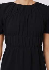 Robe noire avec des manches courtes et bouffantes. Présente un tissu froncé au niveau du décolleté et de la taille, créant une silhouette texturée et fluide.