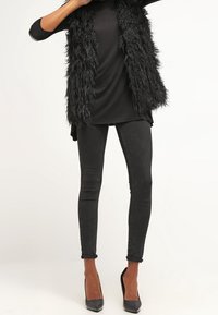 Svart fuskpälsväst över en svart topp, kombinerad med svarta skinny jeans och svarta klackskor. Slät textur och smal passform.