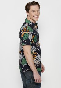 Camisa de manga corta de color azul oscuro con un estampado de hojas tropicales en verde, amarillo y blanco. Presenta un cuello con solapa y un ajuste holgado.