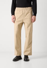 Pantalons beiges à coupe décontractée avec une taille élastique, une coupe droite et une texture lisse, assortis à des chaussures noires.