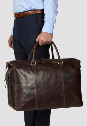 Sac de voyage en cuir marron avec une texture lisse, deux poignées sur le dessus et des accents en métal argenté. Il présente un design à panneaux cousus.
